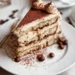 Tiramisu cake topped with mascarpone frosting and cocoa powder, served on a plate.
